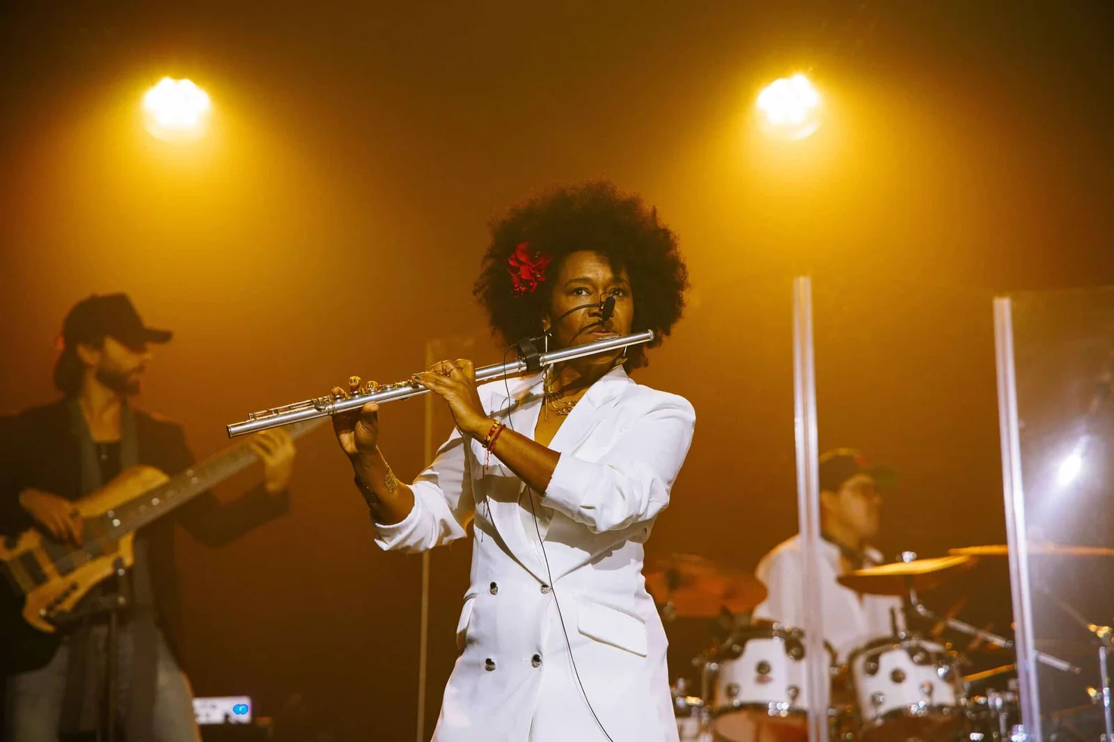 Magela Herrera playing flute in a white jacket with a red flower in her hair, under golden stage lights beside a bassist and drummer
