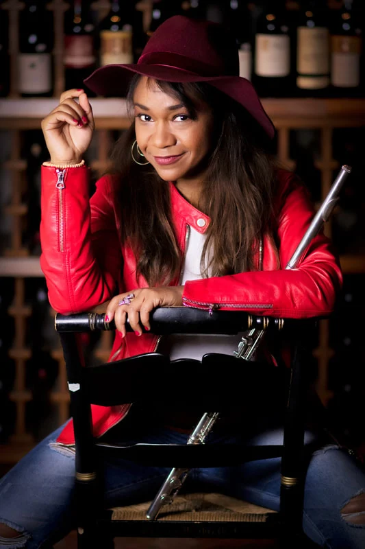 Magela Herrera in a red leather jacket and wine-red hat, seated on a chair holding her flute