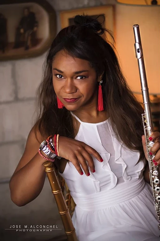 Magela Herrera in a white dress and red tassel earrings, seated and holding her flute