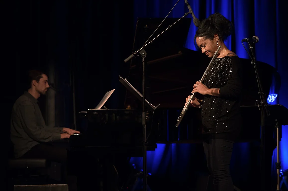 Magela Herrera smiling as she plays flute on stage, a pianist at a grand piano in the background